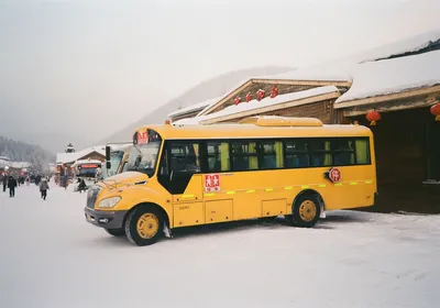 Yellow school bus parked in a snowy village street during winter, surrounded by buildings.